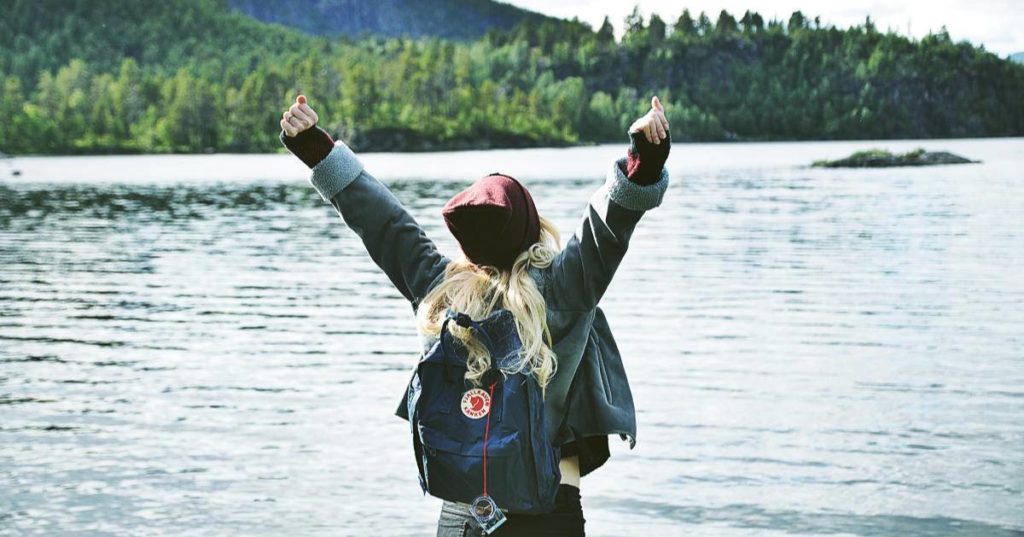 Woman standing by body of water, back to camera, and wearing backpack, with raised hands