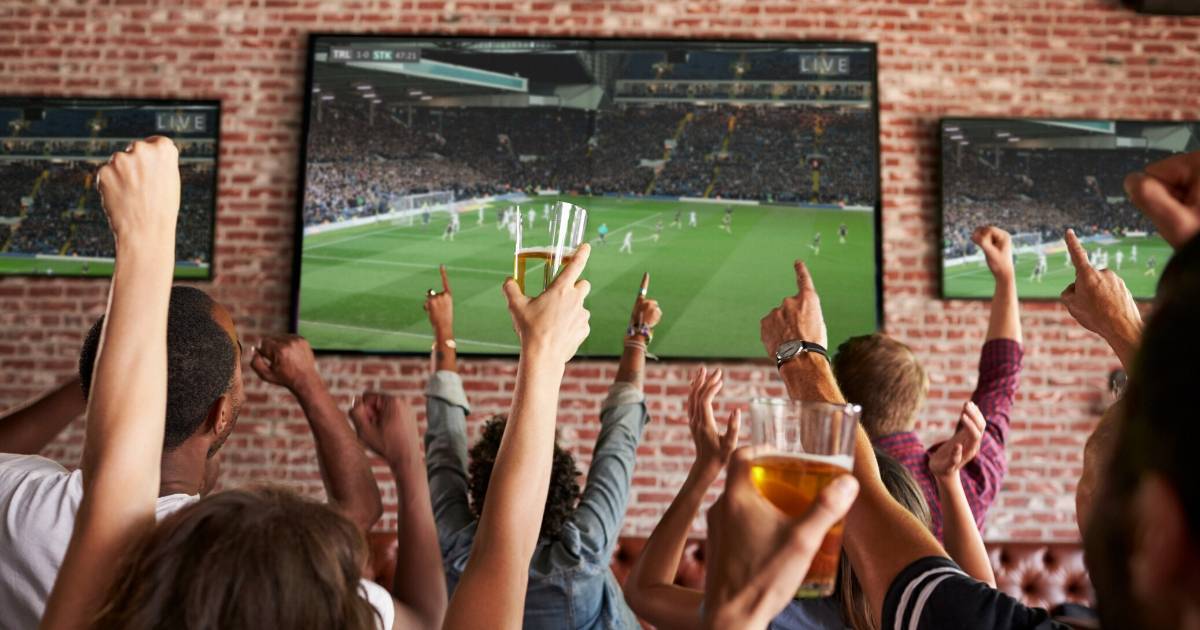 Rear View Of Friends Watching Game In Sports Bar On Screens