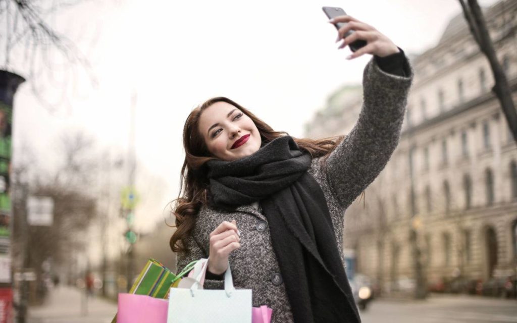 Influencer taking selfie while carrying shopping bags outside. 