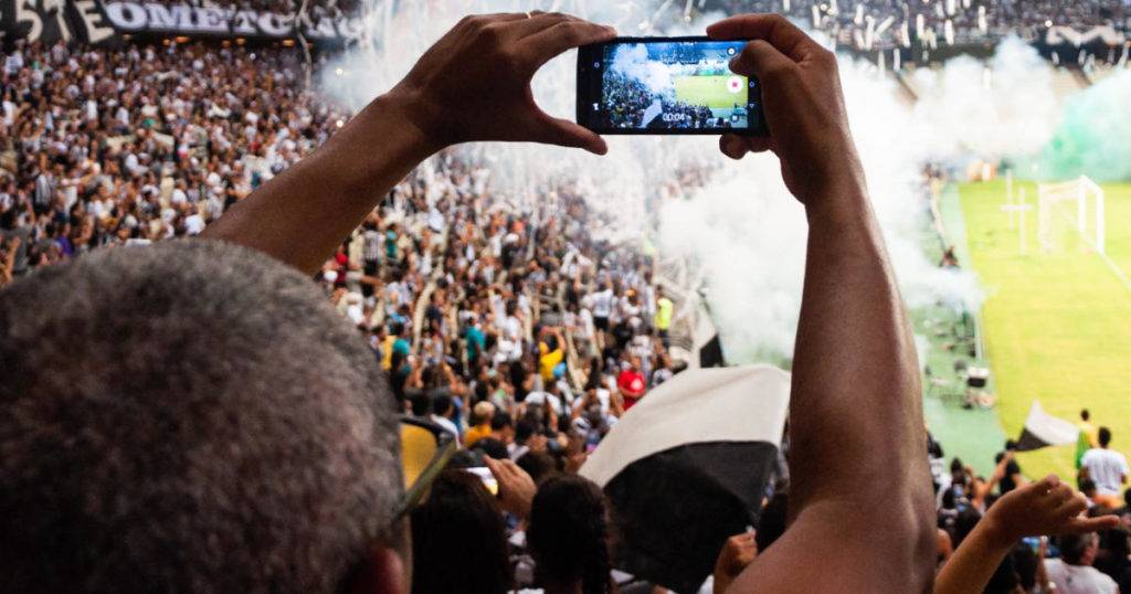 Male fan in crowd holding up mobile phone to take video. Engage gen z sports fans.