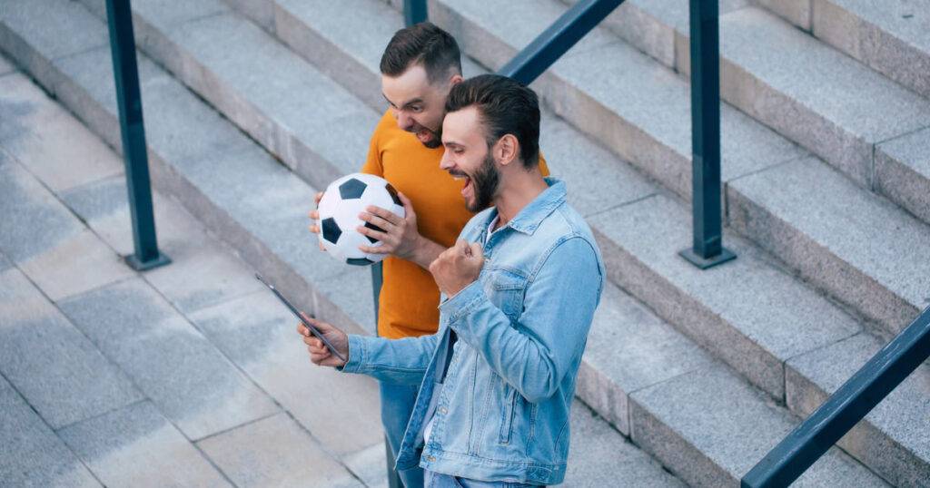 mobile sports app being used by two male sports fans, one holding a soccer ball.
