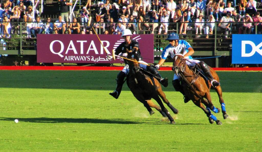 Two polo players on horses in foreground, with sponsor logo signage in background; AI for sports sponsors.